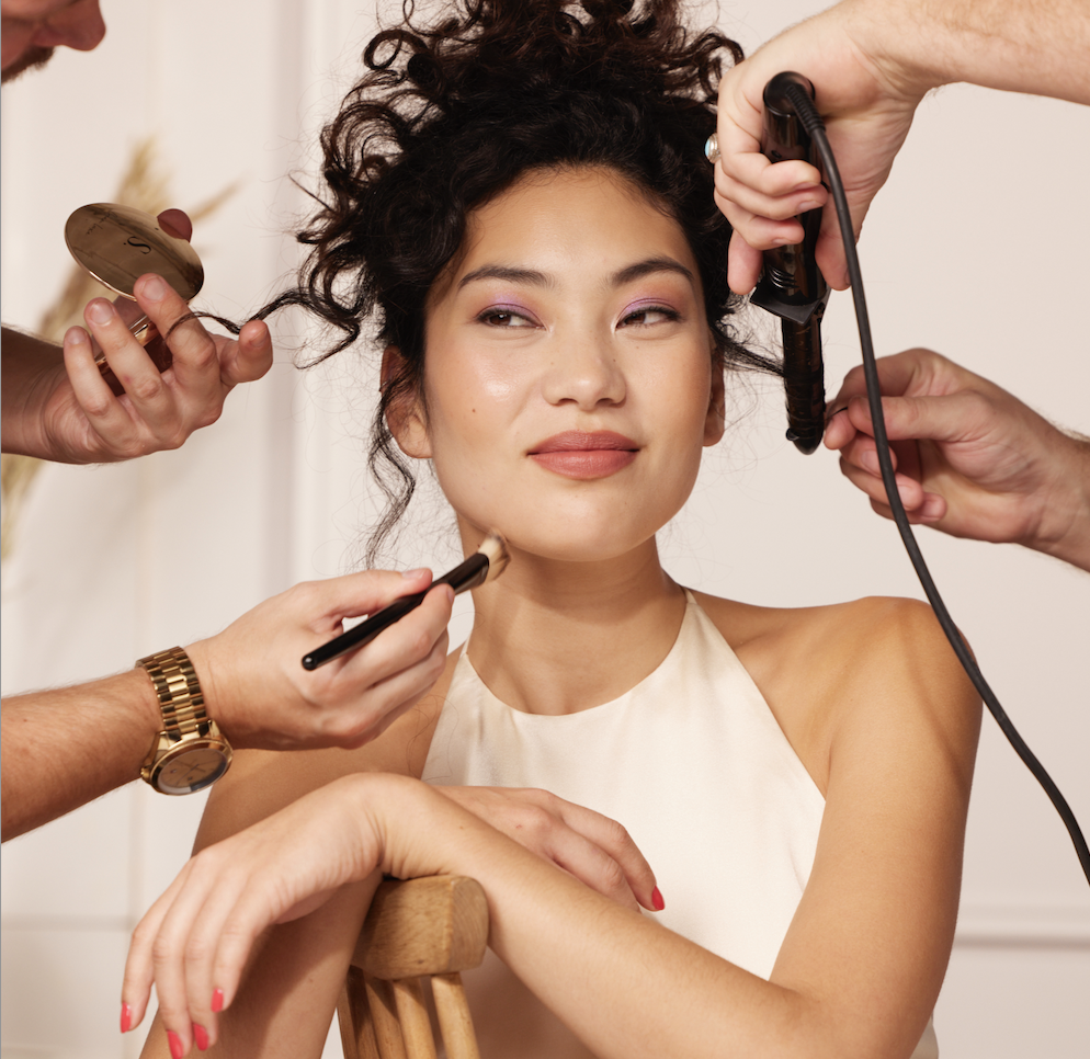 Saks Hair & Beauty collection image featuring a dark brunette model with her curly hair pinned to the back of her head, posing for an image for Saks Hair & Beauty, being prepped by a makeup artist and Luke Pluckrose, creative director for Saks.